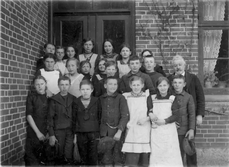 Det ser ud som om at nogle af eleverne fra Ejlstrup Skole smiler. Mon de har fået læst vittighederne fra Dansk Børnetidende højt af lærer Petersen (th)? Billedet er fra 1910. 