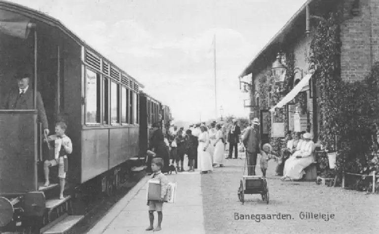 Den gamle jernbanestation i Gilleleje. Gribskovbanen blev forlænget til Gilleleje i 1896. Samme år åbnede det nye Badehotel. Sommergæsterne strømmede nu til byen i større tal. Her på billedet fra 1915 er det før togafgang på Gillelejes første station.