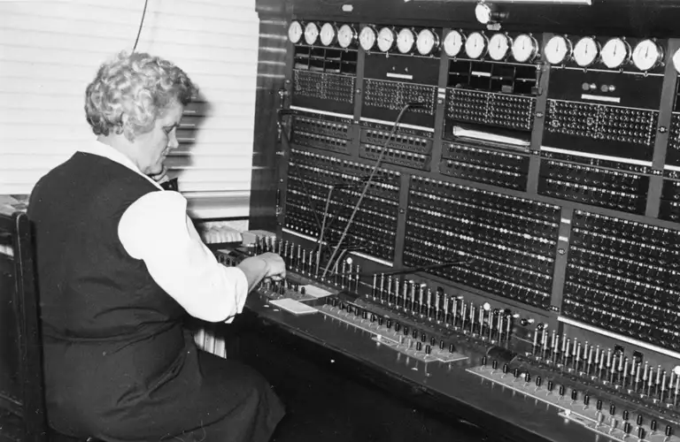 Inger Nielsen ved det manuelle omstillingsbord, ca. 1960’erne. Her forbandt hun de to kabler eller ”propper” med de pågældende abonnenter, så den modtagende abonnents telefon blev ringet op. Foto: Bent Willemoes 